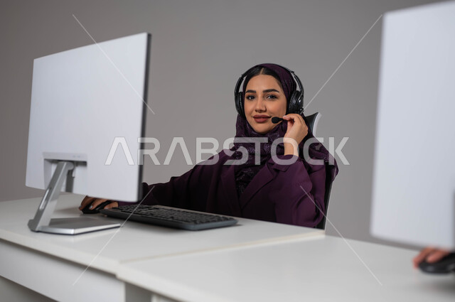 الإجابة على الأسئلة والاستفسارات, قسم الاستعلامات والاتصال، النظر إلى الكاميرا بإيماءات السرور، مفهوم خدمة العملاء، وظائف ومهن سعودية، بورتريه لموظفة عربية خليجية سعودية محجبة مبتسمة تجلس في مقر الشركة ترتدي سماعات الرأس ،خلفية رمادية