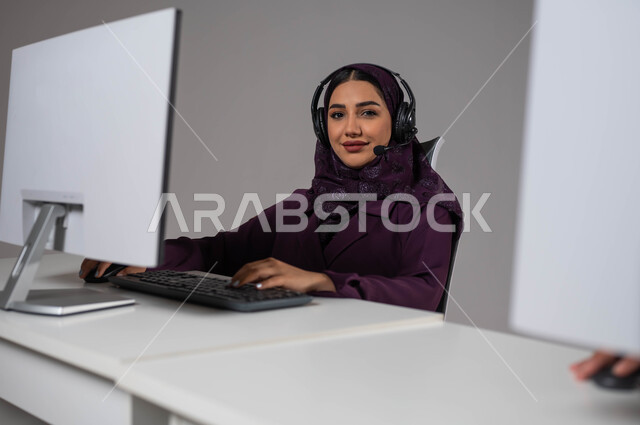 Looking at the camera, wearing a headset to answer questions and inquiries, using modern technologies, attracting and attracting more customers, a portrait of a smiling, veiled Saudi Arabian Gulf woman sitting at the workplace and working in technical support and customer service, gray background