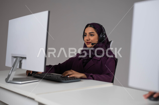 ارتداء سماعة الاتصال للإجابة عن الاستعلامات والاستفسارات ،زيادة مستوى رضا العميل, استخدام التقنيات الحديثة، استقطاب و جذب المزيد من العملاء، بورتريه لسيدة عربيه سعوديه خليجيه محجبة تجلس في مقر العمل و تعمل في الدعم الفني وخدمة العملاء, خلفية رمادية