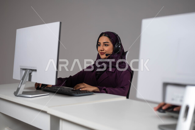 Increasing the level of customer satisfaction, using modern technologies, attracting more customers, a portrait of a veiled Saudi Arabian Gulf woman sitting at the workplace and working in technical support and customer service, wearing a communication headset to answer inquiries and questions, gray background
