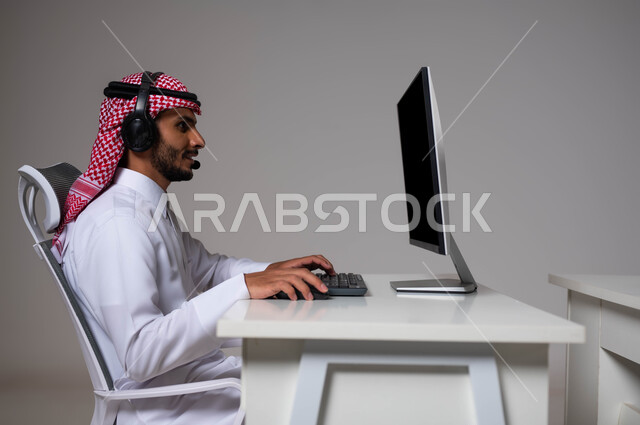 ارتداء سماعات الاتصال ،دمج التقنية والتكنولوجيا مع الحياة العملية, مفهوم خدمة العملاء، بورتريه مقرب لموظف عربي خليجي سعودي يجلس في مقر الشركة يرتدي الشماغ والثوب التقليدي يقوم بالإجابة على الاسئلة والاستفسارات عبر جهاز الحاسوب, خلفية رمادية.
