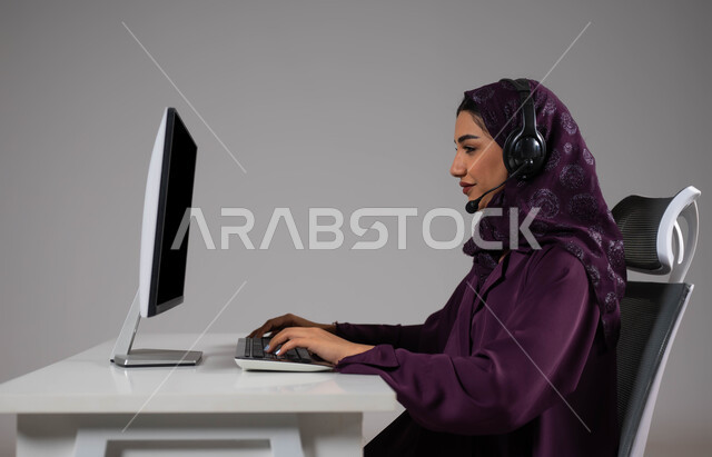 استخدام التقنيات الحديثة، استقطاب و جذب المزيد من العملاء، العمل على زيادة مستوى رضا العميل، الإجابة عن الاستعلامات والاستفسارات, بورتريه مقرب لسيدة عربيه سعوديه خليجيه محجبة مبتسمة تجلس على كرسي مريح ترتدي سماعة الرأس و تعمل على جهاز الحاسوب, خلفية رمادية