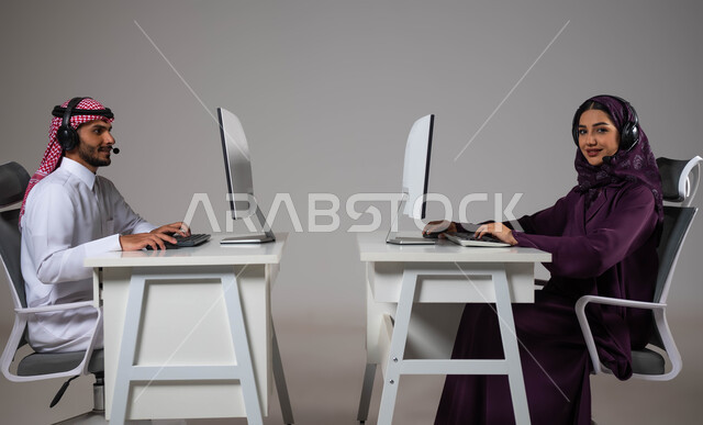 ارتداء سماعة الاتصال للتواصل مع العملاء ،الإجابة على الاستعلامات والاستفسارات, بورتريه لرجل و امرأة عربيان سعوديان خليجيان يعملان في خدمة العملاء ويجلسون في مقر العمل ،متابعة وتطوير العمل في الشركات السعودية ،مهن ووظائف سعودية، خلفية رمادية .