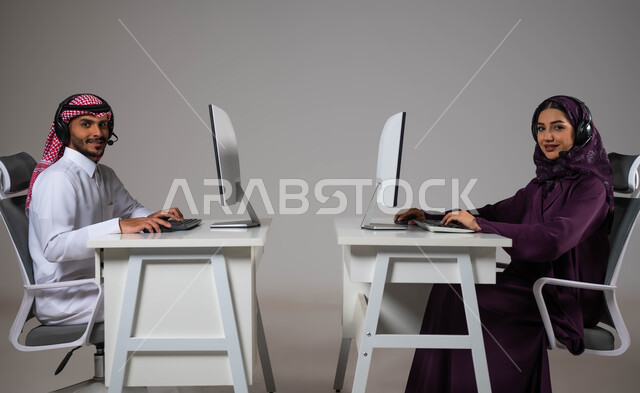 Answering questions and inquiries, a portrait of two Saudi Gulf Arab employees working in customer service looking at the camera wearing a communication headset, working to increase the level of customer satisfaction, Saudi professions and jobs, gray background