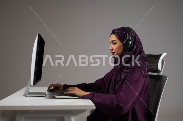 العمل على زيادة مستوى رضا العميل، جذب واستقطاب المزيد من العملاء، الإجابة عن الاستعلامات والاستفسارات, بورتريه لسيدة عربيه سعوديه خليجيه محجبة مبتسمة تجلس على كرسي مريح تنظر إلى الكاميرا ترتدي سماعة الرأس تعمل على جهاز الحاسوب, خلفية رمادية .