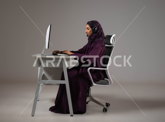 التركيز على مستوى رضا العميل، العمل على جذب واستقطاب المزيد من العملاء، الإجابة عن الاستعلامات والاستفسارات, بورتريه لسيدة عربيه سعوديه خليجيه محجبة مبتسمة تجلس على كرسي مريح تنظر إلى الكاميرا ترتدي سماعة الرأس تعمل على جهاز الحاسوب, خلفية رمادية .