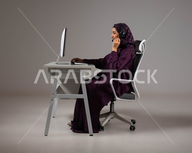 العمل على جذب واستقطاب المزيد من العملاء، تعزيز مستوى رضا العميل، الإجابة عن الاستعلامات والاستفسارات, بورتريه لسيدة عربيه سعوديه خليجيه محجبة مبتسمة تجلس على كرسي مريح تنظر إلى الكاميرا ترتدي سماعة الرأس تعمل على جهاز الحاسوب, خلفية رمادية .