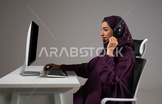 Meeting the needs of the public, enhancing the level of customer satisfaction, answering queries and inquiries, a portrait of a smiling, veiled Saudi Arabian Gulf woman sitting in a comfortable chair looking at the camera wearing a headset working on the computer, working in customer service, gray background