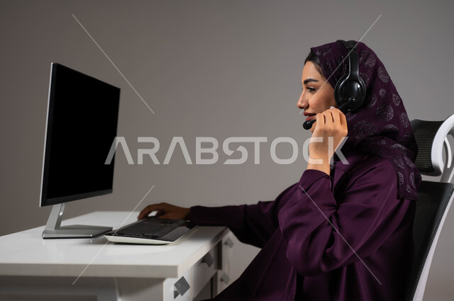 احتراف مهارات التواصل الفعال، تعزيز مستوى رضا العميل، الإجابة عن الاستعلامات والاستفسارات, بورتريه لسيدة عربيه سعوديه خليجيه محجبة مبتسمة تجلس على كرسي مريح تنظر إلى الكاميرا ترتدي سماعة الرأس تعمل على جهاز الحاسوب, العمل في خدمة العملاء، خلفية رمادية .