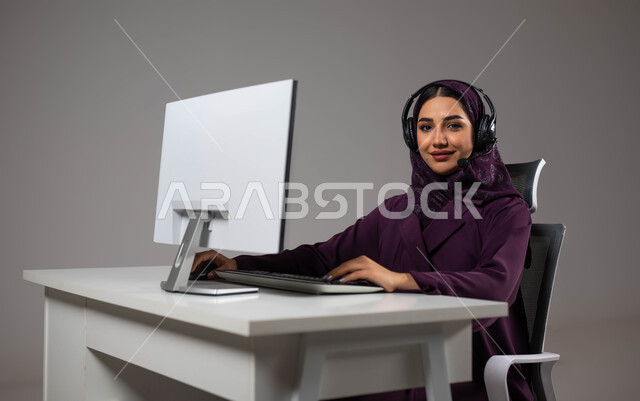 مهارات التواصل الفعال، تعزيز مستوى رضا العميل، الإجابة عن الاستعلامات والاستفسارات, بورتريه لسيدة عربيه سعوديه خليجيه محجبة مبتسمة تجلس على كرسي مريح تنظر إلى الكاميرا ترتدي سماعة الرأس تعمل على جهاز الحاسوب, العمل في خدمة العملاء، خلفية رمادية .