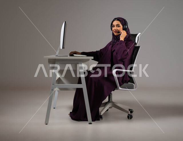 Listening and listening skills, enhancing the level of customer satisfaction, answering queries and enquiries, a portrait of a smiling, veiled Saudi Arabian Gulf woman sitting in a comfortable chair looking at the camera wearing a headset working on the computer, working in customer service, gray background.