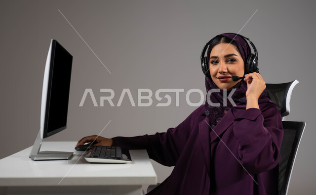 تعابير وإيماءات الثقة بالنفس، تعزيز مستوى رضا العميل، الإجابة عن الاستعلامات والاستفسارات, بورتريه لسيدة عربيه سعوديه خليجيه محجبة مبتسمة تجلس على كرسي مريح تنظر إلى الكاميرا ترتدي سماعة الرأس تعمل على جهاز الحاسوب, العمل في خدمة العملاء، خلفية رمادية .
