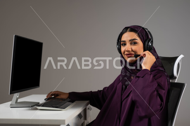تعزيز مستوى رضا العميل، النظر إلى الكاميرا بإيماءات الثقة، الإجابة عن الاستعلامات والاستفسارات, بورتريه لسيدة عربيه سعوديه خليجيه محجبة مبتسمة تجلس على كرسي مريح ترتدي سماعة الرأس تعمل على جهاز الحاسوب, العمل في خدمة العملاء، خلفية رمادية .