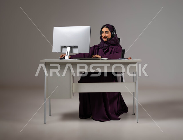 إنجاز المهام المكتبية، الإجابة عن الاستعلامات والاستفسارات, احتراف وظائف الحاسب والتكنولوجيا، بورتريه لإمراه عربيه سعوديه خليجيه محجبة مبتسمة تجلس على كرسي مريح ترتدي سماعة الرأس تعمل على جهاز الحاسوب,  العمل في خدمة العملاء، خلفية رمادية .