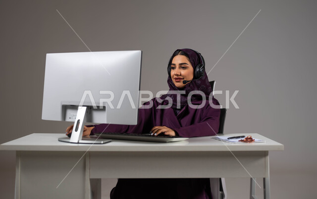 الإجابة عن الاستعلامات والاستفسارات, احتراف وظائف الحاسب والتكنولوجيا، بورتريه لإمراه عربيه سعوديه خليجيه محجبة مبتسمة تجلس على كرسي مريح ترتدي سماعة الرأس تعمل على جهاز الحاسوب,  العمل في خدمة العملاء، إنجاز المهام المكتبية، خلفية رمادية .