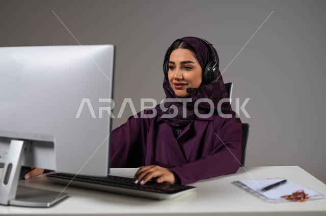احتراف وظائف الحاسب والتكنولوجيا، بورتريه لإمراه عربيه سعوديه خليجيه محجبة مبتسمة تجلس على كرسي مريح ترتدي سماعة الرأس تعمل على جهاز الحاسوب,  العمل في خدمة العملاء، الإجابة عن الاستعلامات والاستفسارات, إنجاز المهام المكتبية، خلفية رمادية .