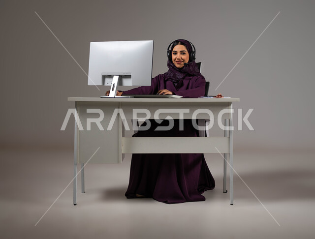النظر إلى الكاميرا بإيماءات السرور، مفهوم خدمة العملاء، وظائف ومهن سعودية، تطوير استراتيجية العمل، بورتريه مقرب لموظفة عربية خليجية سعودية محجبة مبتسمة تجلس على كرسي مريح ترتدي سماعات الرأس تقوم بالإجابة على الأسئلة والاستفسارات, خلفية رمادية.
