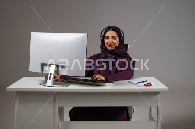 قسم الاستعلامات والاتصال، النظر إلى الكاميرا بإيماءات السرور، مفهوم خدمة العملاء، وظائف ومهن سعودية،  بورتريه مقرب لموظفة عربية خليجية سعودية محجبة مبتسمة تجلس على كرسي مريح ترتدي سماعات الرأس تقوم بالإجابة على الأسئلة والاستفسارات, خلفية رمادية.