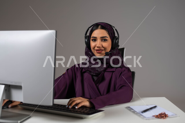مهن ووظائف سعودية، تطوير استراتيجية العمل، مفهوم خدمة العملاء، دمج العمل مع التقنية والتكنولوجيا, بورتريه لموظفة عربية خليجية سعودية تجلس على كرسي مريح ترتدي سماعات الرأس تقوم بالإجابة على الأسئلة والاستفسارات, خلفية رمادية.