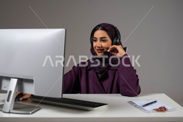 الاندماج والتركيز في آداء مهام العمل، مفهوم خدمة العملاء، وظائف ومهن سعودية، تطوير استراتيجية العمل، بورتريه مقرب لموظفة عربية خليجية سعودية محجبة مبتسمة تجلس على كرسي مريح ترتدي سماعات الرأس تقوم بالإجابة على الأسئلة والاستفسارات, خلفية رمادية.