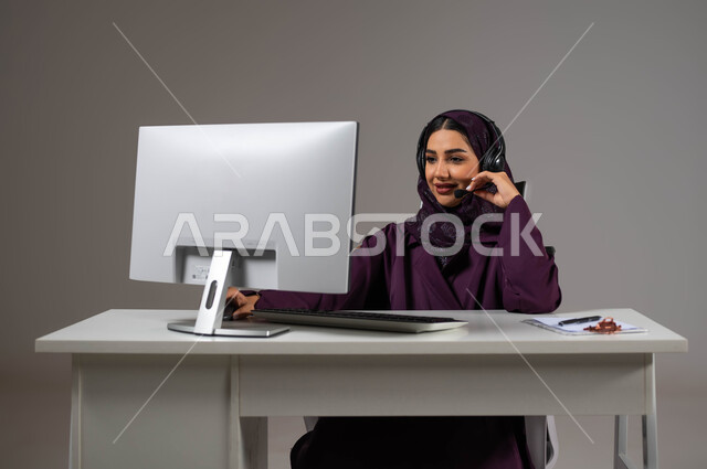 وظائف ومهن سعودية، تطوير استراتيجية العمل، مفهوم خدمة العملاء، دمج العمل مع التقنية والتكنولوجيا, بورتريه مقرب لموظفة عربية خليجية سعودية محجبة مبتسمة تجلس على كرسي مريح ترتدي سماعات الرأس تقوم بالإجابة على الأسئلة والاستفسارات, خلفية رمادية.