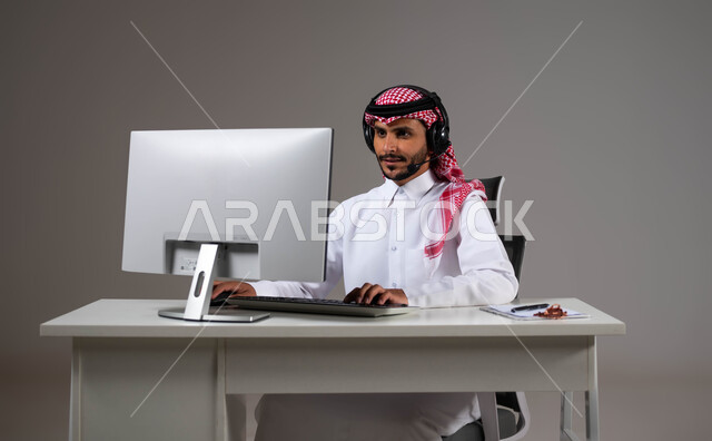 التركيز والتمعن أثناء إنجاز مهام العمل، مفهوم خدمة العملاء، دمج العمل مع التقنية والتكنولوجيا, بورتريه مقرب لموظف عربي خليجي سعودي يجلس على كرسي مريح يرتدي الشماغ والثوب التقليدي وسماعات الرأس ينظر إلى الكاميرا بإيماءات الثقة, خلفية رمادية.