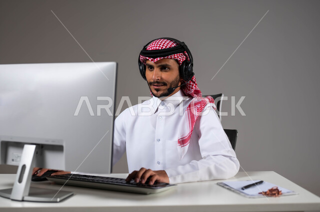 إيماءات التركيز لإنجاز مهام العمل، مفهوم خدمة العملاء، دمج العمل مع التقنية والتكنولوجيا, بورتريه مقرب لموظف عربي خليجي سعودي يجلس على كرسي مريح يرتدي الشماغ والثوب التقليدي وسماعات الرأس ينظر إلى الكاميرا بإيماءات الثقة, خلفية رمادية.