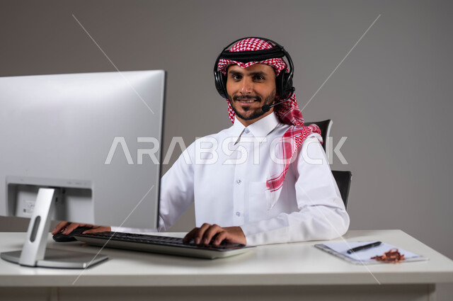 استخدام الحاسوب المحمول لإنجاز المهام، مفهوم خدمة العملاء، دمج العمل مع التقنية والتكنولوجيا, بورتريه مقرب لموظف عربي خليجي سعودي يجلس على كرسي مريح يرتدي الشماغ والثوب التقليدي وسماعات الرأس ينظر إلى الكاميرا بإيماءات الثقة, خلفية رمادية.