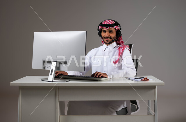النظر إلى الكاميرا بإيماءات الثقة بالنفس، مفهوم خدمة العملاء، بورتريه لرجل عربي خليجي سعودي يجلس على كرسي مريح يرتدي الشماغ والثوب التقليدي وسماعات الرأس يقوم بالإجابة على الاسئلة والاستفسارات عبر جهاز الحاسوب, خلفية رمادية.