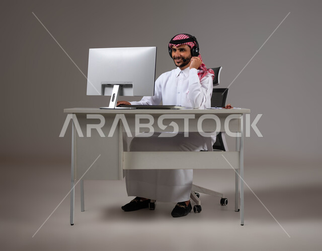 إيماءات التمعن والتركيز، استخدام التقنية والتكنولوجيا الحديثة, مفهوم خدمة العملاء، بورتريه لرجل عربي خليجي سعودي يجلس على كرسي مريح يرتدي الشماغ والثوب التقليدي وسماعات الرأس يقوم بالإجابة على الاسئلة والاستفسارات عبر جهاز الحاسوب, خلفية رمادية.