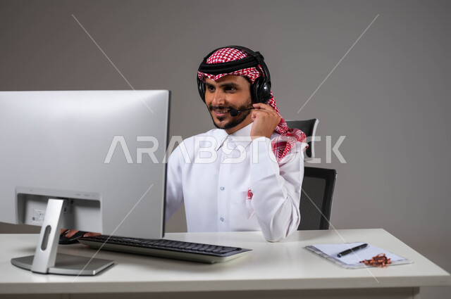 استخدام التقنية والتكنولوجيا الحديثة, مفهوم خدمة العملاء، بورتريه مقرب لموظف عربي خليجي سعودي يجلس على كرسي مريح يرتدي الشماغ والثوب التقليدي وسماعات الرأس يقوم بالإجابة على الاسئلة والاستفسارات عبر جهاز الحاسوب, خلفية رمادية.