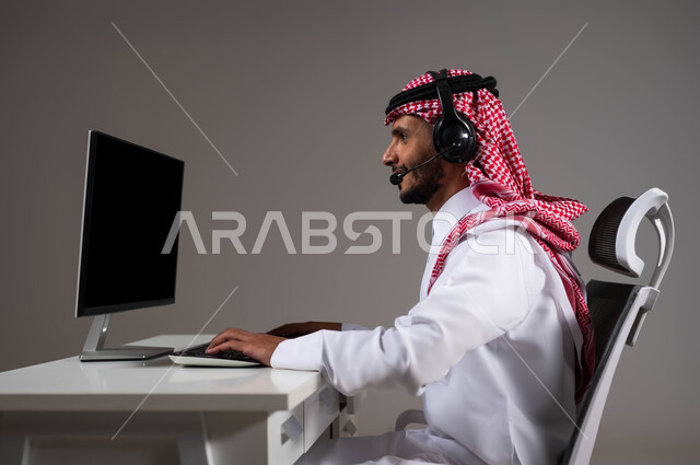 مفهوم خدمة العملاء، دمج العمل مع التقنية والتكنولوجيا, بورتريه مقرب لموظف عربي خليجي سعودي يجلس على كرسي مريح يرتدي الشماغ والثوب التقليدي وسماعات الرأس يقوم بالإجابة على الاسئلة والاستفسارات عبر جهاز الحاسوب, خلفية رمادية.