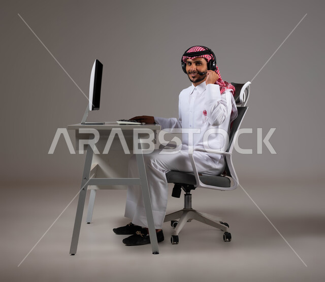 الإجابة على الاسئلة والاستفسارات، مفهوم دمج العمل مع التقنية والتكنولوجيا, شركات ووظائف سعودية, بورتريه مقرب لموظف عربي خليجي سعودي يجلس على كرسي مريح يرتدي الشماغ والثوب التقليدي وسماعات الرأس يقوم بإنجاز مهام العمل باستخدام جهاز الحاسوب, خلفية رمادية.