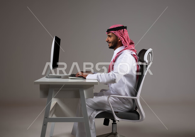 مفهوم دمج العمل مع التقنية والتكنولوجيا, إيماءات التركيز لتحقيق أهداف الشركة، شركات ووظائف سعودية, بورتريه مقرب لموظف عربي خليجي سعودي يجلس على كرسي مريح يرتدي الشماغ والثوب التقليدي يقوم بإنجاز مهام العمل باستخدام جهاز الحاسوب, خلفية رمادية.
