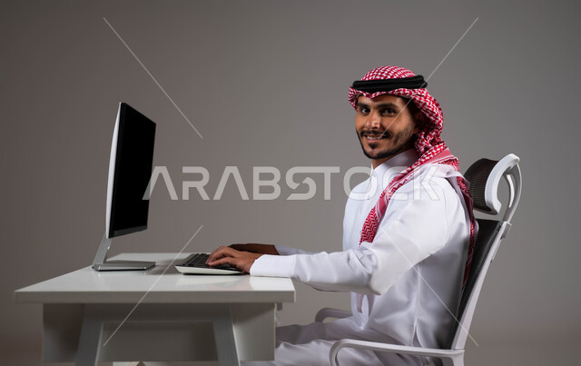 تحقيق أهداف الشركة، مفهوم دمج العمل مع التقنية والتكنولوجيا, بورتريه مقرب لرجل عربي خليجي سعودي يجلس على كرسي مريح يرتدي الشماغ والثوب التقليدي يقوم بإنجاز مهام العمل باستخدام جهاز الحاسوب, النظر إلى الكاميرا بإيماءات الثقة بالنفس، خلفية رمادية.