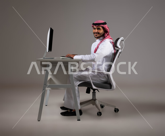 النظر إلى الكاميرا بإيماءات الثقة بالنفس، تحقيق أهداف الشركة، مفهوم دمج العمل مع التقنية والتكنولوجيا, بورتريه مقرب لرجل عربي خليجي سعودي يجلس على كرسي مريح يرتدي الشماغ والثوب التقليدي يقوم بإنجاز مهام العمل باستخدام جهاز الحاسوب, خلفية رمادية.