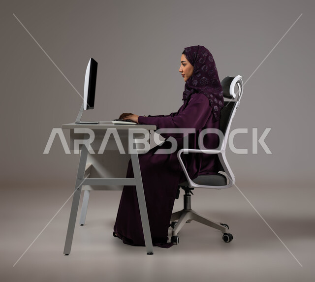 استخدام التقنيات والتكنولوجيا الحديثة، تحقيق أهداف الشركة، إنجاز مهام العمل اليومية، مناقشة وشرح الأعمال عن بعد، بورتريه لسيدة عربية خليجية سعودية محجبة تجلس على كرسي مريح تعمل على جهاز الحاسوب، خلفية رمادية .