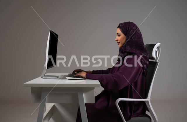 تحقيق أهداف الشركة، إنجاز مهام العمل اليومية، مناقشة وشرح الأعمال عن بعد، بورتريه مقرب لموظفة عربية خليجية سعودية محجبة تجلس على كرسي مريح تعمل على جهاز الحاسوب، استخدام تقنيات التكنولوجيا الحديثة، خلفية رمادية .