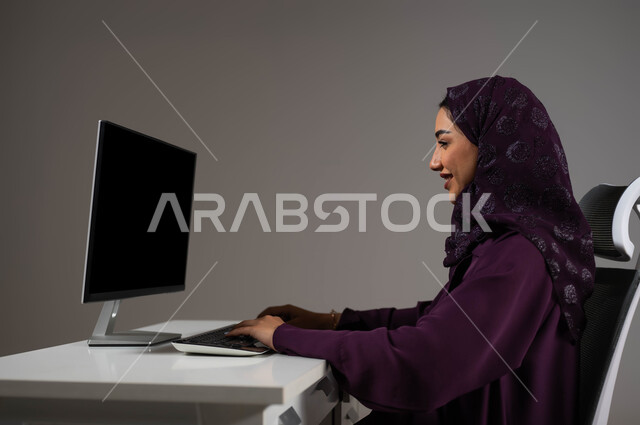 إنجاز مهام العمل اليومية، تحقيق أهداف الشركة، مناقشة وشرح الأعمال عن بعد، بورتريه مقرب لموظفة عربية خليجية سعودية محجبة تجلس على كرسي مريح تعمل على جهاز الحاسوب، استخدام تقنيات التكنولوجيا الحديثة، خلفية رمادية .