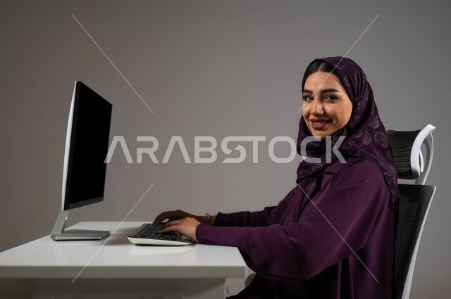 العمل في خدمة العملاء، استخدام التقنيات الحديثة والمتطورة، بورتريه مقرب لموظفة عربية خليجية سعودية مبتسمة تنظر إلى الكاميرا بإيماءات الثقة بالنفس، استعلامات واستفسارات والاجابه على اسئلة الزبائن، اجراء اتصال فيديو من خلال الحاسوب، خلفية رمادية .