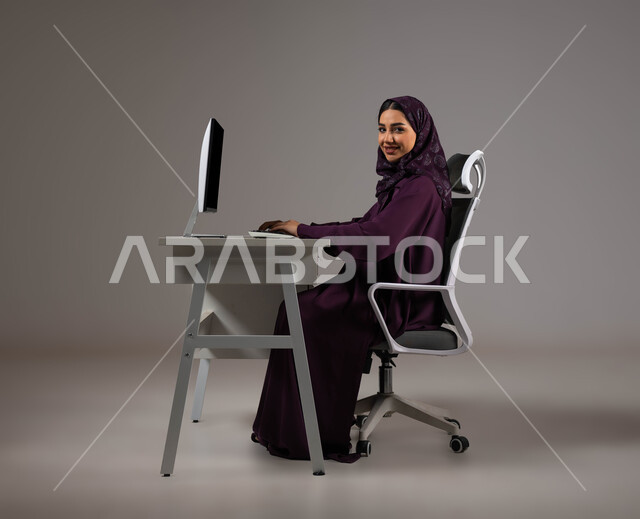 استخدام التقنيات الحديثة والمتطورة، بورتريه لموظفة عربية خليجية سعودية مبتسمة تنظر إلى الكاميرا بإيماءات الثقة بالنفس، العمل في خدمة العملاء، استعلامات واستفسارات والاجابه على اسئلة الزبائن، اجراء اتصال فيديو من خلال الحاسوب، خلفية رمادية .