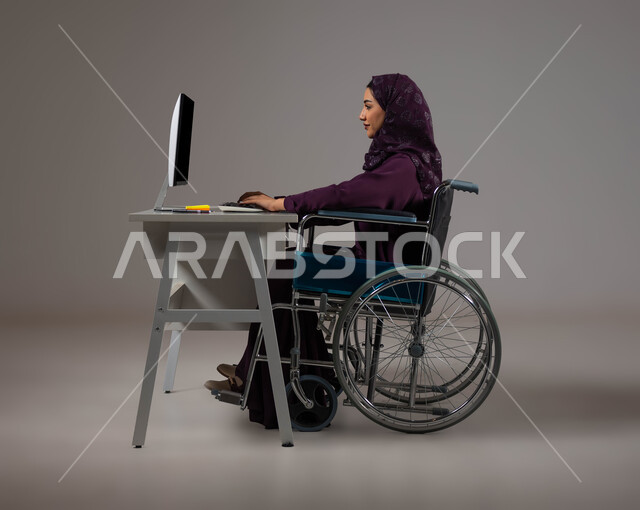 توظيف ودمج ذوي الإعاقة في مختلف المجالات، إتاحة المجال امام ذوي الهمم للدخول الى مجال العمل، التمكين الاجتماعي في المجتمع السعودي، بورتريه لإمرأة عربية خليجية سعودية محجبة تجلس على الكرسي المتحرك تعمل على جهاز الحاسوب في مقر الشركة,  خلفية رمادية