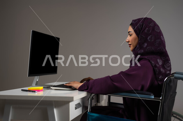 إتاحة المجال امام ذوي الهمم للدخول الى مجال العمل، التمكين الاجتماعي في المجتمع السعودي، توفير بيئة عمل مريحة للمرضى والمعاقين، بورتريه لإمرأة عربية خليجية سعودية محجبة تجلس على الكرسي المتحرك تعمل على جهاز الحاسوب في مقر الشركة,  خلفية رمادية