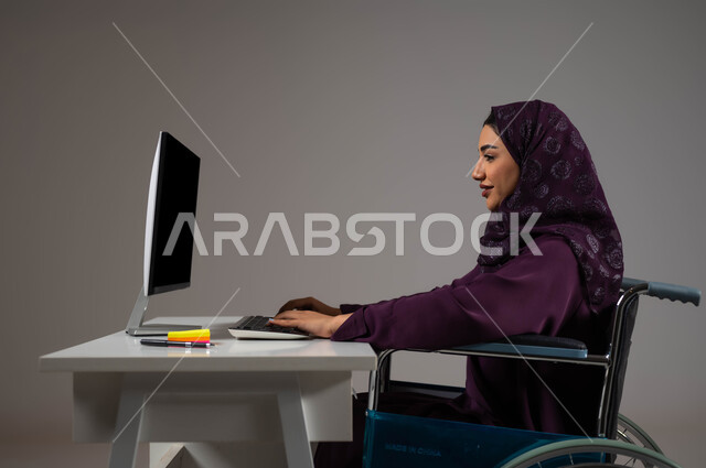 توفير بيئة عمل مريحة للمرضى والمعاقين، التمكين الاجتماعي في المجتمع السعودي، بورتريه مقرب لإمرأة عربية خليجية سعودية محجبة تجلس على الكرسي المتحرك تعمل على جهاز الحاسوب في مقر الشركة, إتاحة المجال امام ذوي الهمم للدخول الى مجال العمل، خلفية رمادية