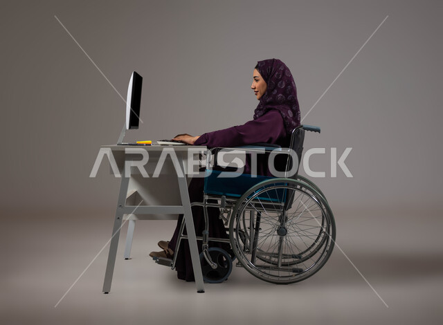 التمكين الاجتماعي في المجتمع السعودي، توفير بيئة عمل مريحة للمرضى والمعاقين، بورتريه لإمرأة عربية خليجية سعودية محجبة تجلس على الكرسي المتحرك تعمل على جهاز الحاسوب في مقر الشركة, إتاحة المجال امام ذوي الهمم للدخول الى مجال العمل، خلفية رمادية