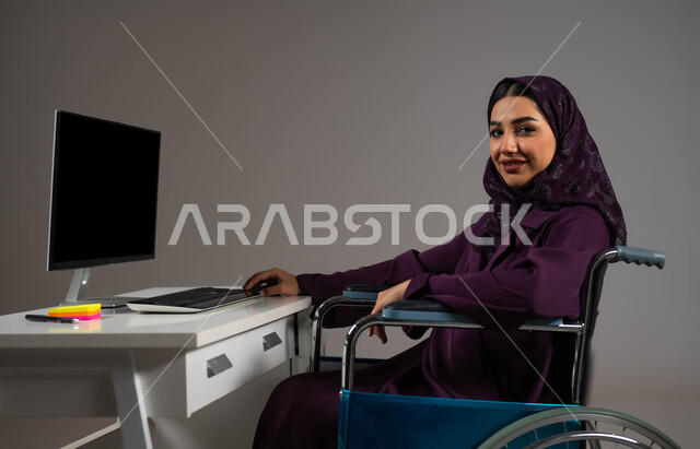 الشعور بالراحه والطمأنينه، اهتمام ودعم المجتمع السعودي لذوي الاحتياجات الخاصه, دمج و توظيف ذوي الاعاقه في مختلف المجالات, بورتريه مقرب لإمرأة عربية سعودية خليجية محجبة مبتسمة من ذوي الهمم تنظر إلى الكاميرا تجلس في مقر العمل, خلفية رمادية .