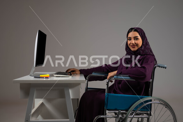 اهتمام ودعم المجتمع السعودي لذوي الاحتياجات الخاصه, دمج و توظيف ذوي الاعاقه في مختلف المجالات, بورتريه مقرب لإمرأة عربية سعودية خليجية محجبة مبتسمة من ذوي الهمم تنظر إلى الكاميرا تجلس في مقر العمل, الشعور بالراحه والطمأنينه، خلفية رمادية .