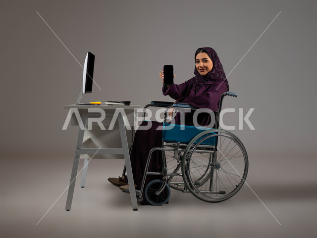 الشعور بالراحه والطمأنينه، دمج و توظيف ذوي الاعاقه في مختلف المجالات, بورتريه لموظفة عربية سعودية خليجية محجبة مبتسمة من ذوي الهمم تنظر إلى الكاميرا تعرض الهاتف المحمول بشاشة سوداء فارغة, اهتمام  ودعم المجتمع السعودي لذوي الاحتياجات الخاصه, خلفية رمادية .