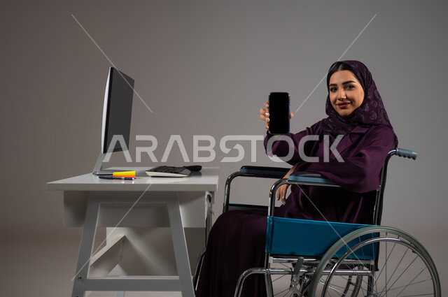 Saudi society’s support and interest in people with special needs, integration and employment of people with disabilities in various fields, feeling of comfort and reassurance, portrait of a smiling, veiled Saudi Arabian Gulf employee with special needs looking at the camera showing the mobile phone with a blank black screen, gray background