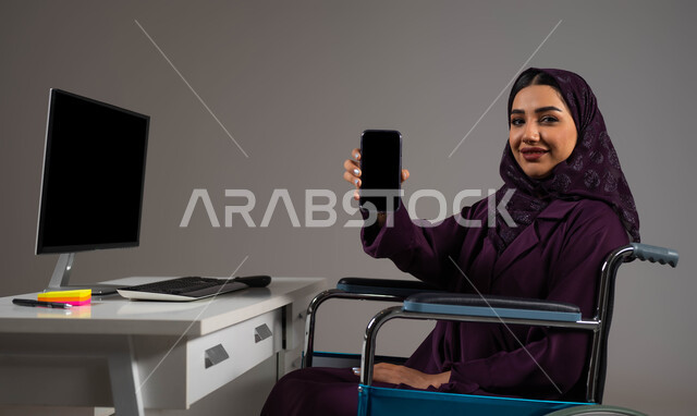 فتح المجال امام ذوي الهمم للدخول الى مجال العمل, تسهيل حياة المعاقين باستخدام التقنية والتكنولوجيا, بورتريه مقرب لشابة عربية خليجية سعودية مبتسمة ترتدي العباءة والحجاب تجلس على كرسي متحرك تقوم بعرض شاشة سوداء فارغه على الهاتف المحمول داخل المكتب, خلفية رمادية